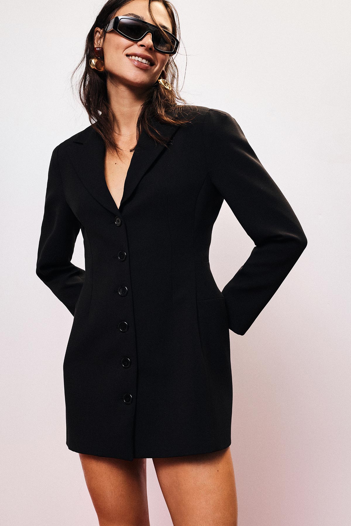 Smiling woman in black tailored blazer dress with sunglasses and gold hoop earrings, hands in pockets, studio background