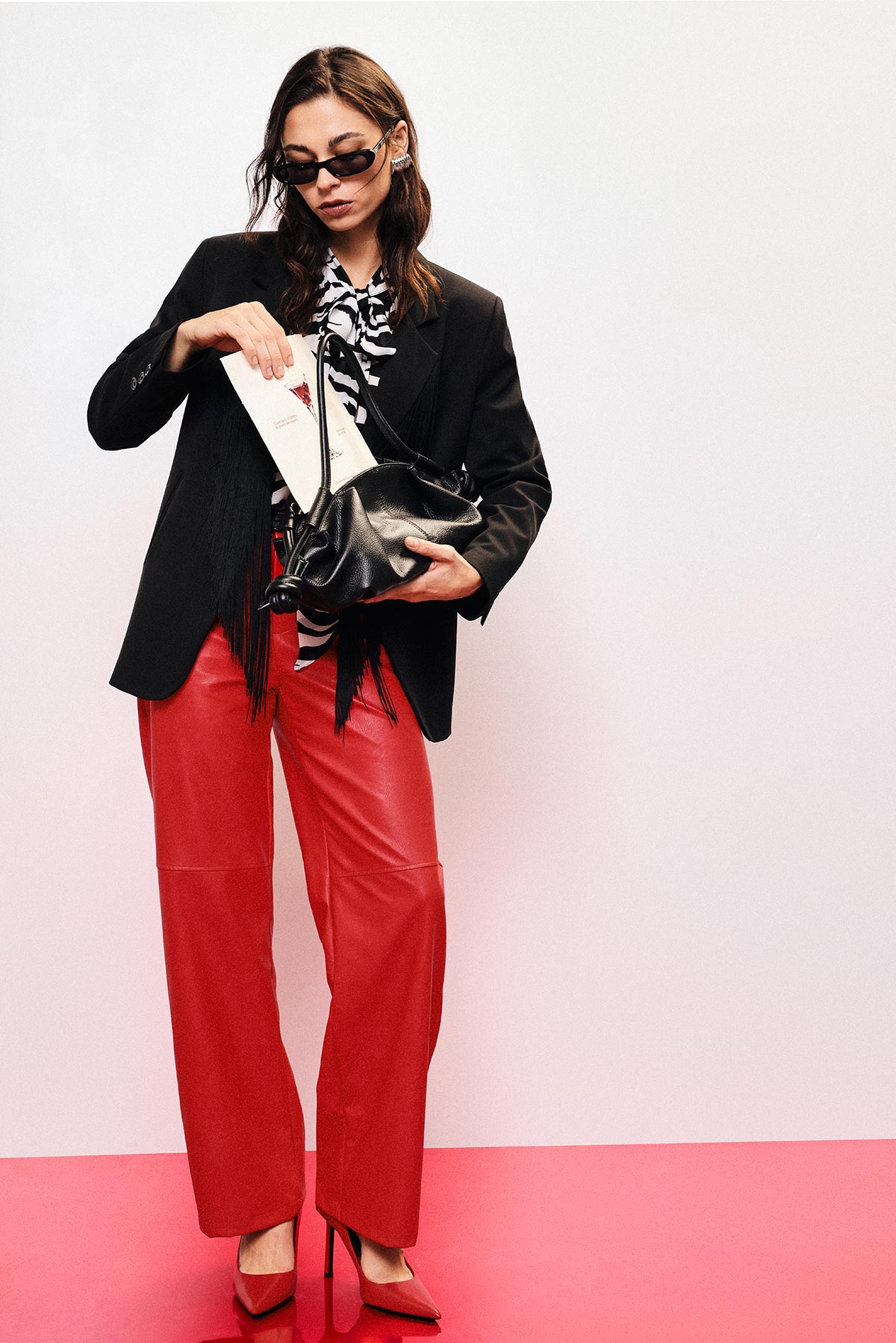 Fashion model in sunglasses wearing black blazer, zebra blouse and red leather trousers, holding black handbag and envelope, red heels