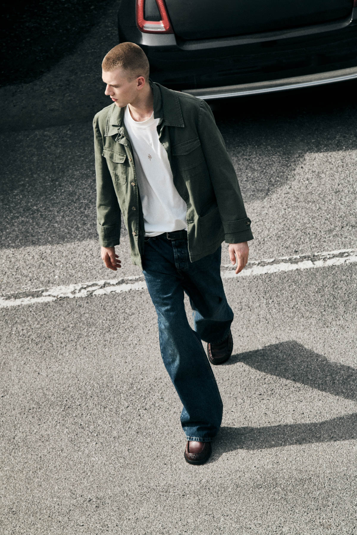 Young man in green jacket and white T‑shirt walking past a parked car on tarmac, looking left