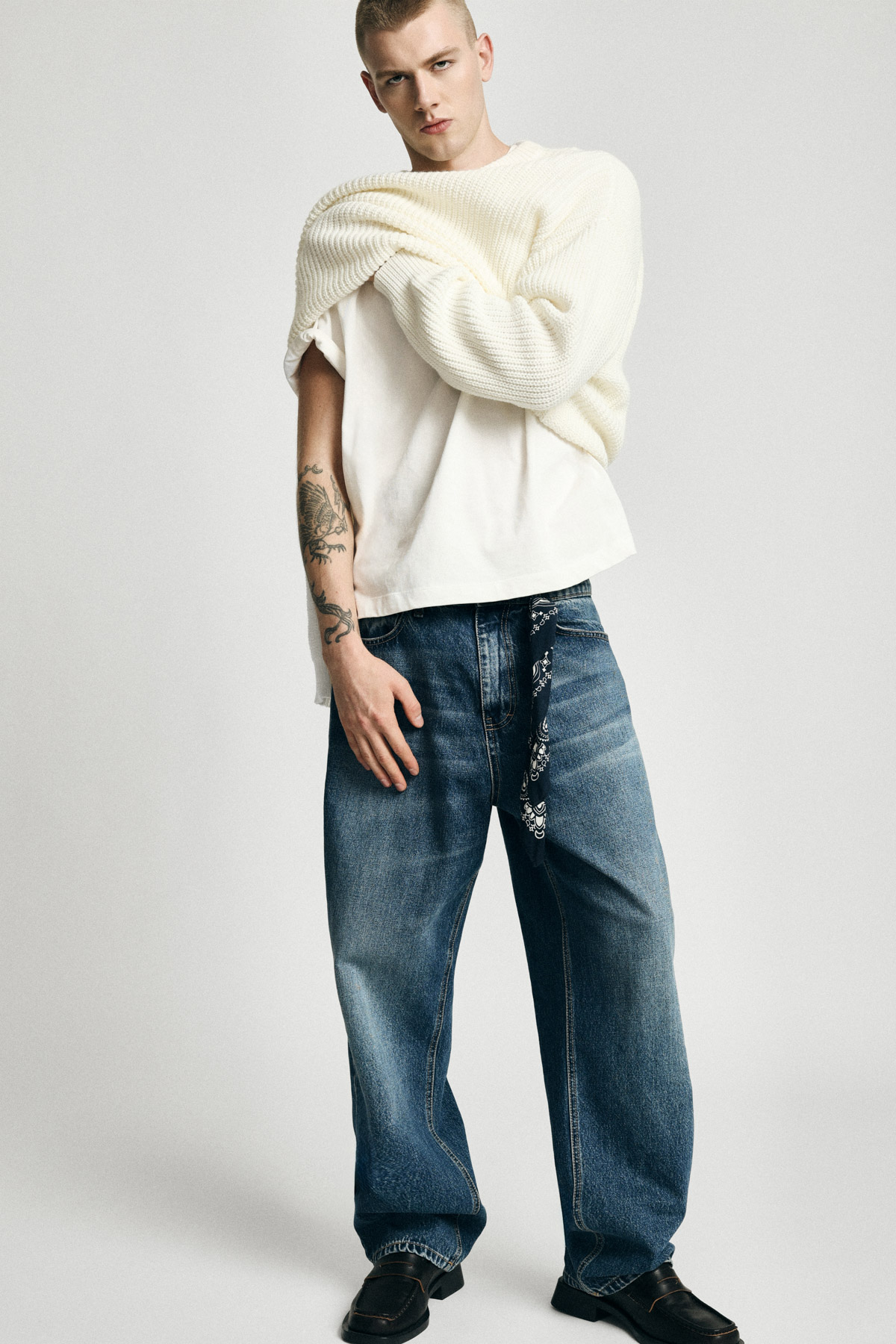 Male model wearing cream knit sweater over white tee, baggy blue jeans with bandana belt, forearm tattoos and black shoes