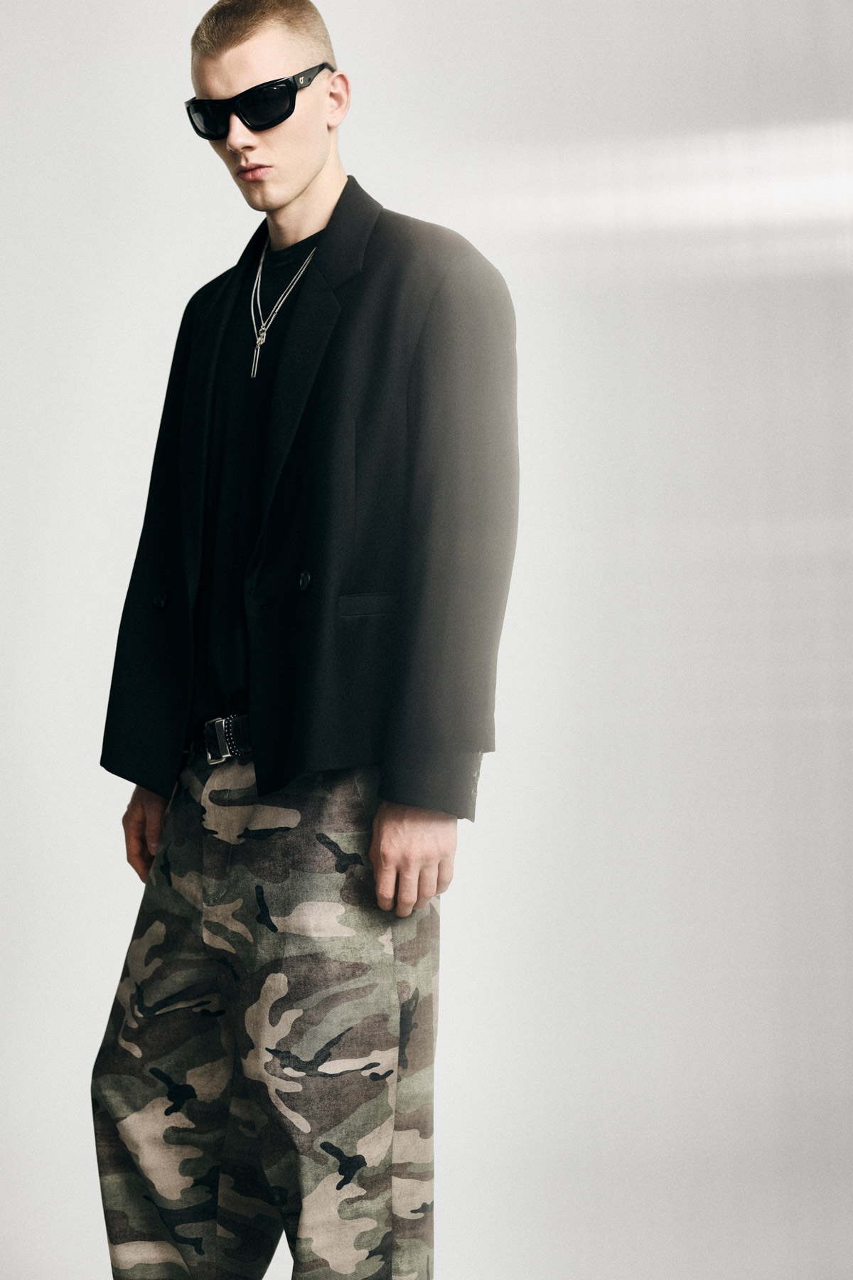 Male model in black blazer, sunglasses and layered necklaces wearing camouflage trousers in a minimalist studio fashion shot