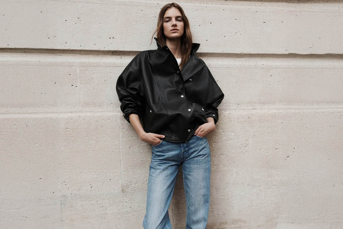 Model wearing oversized black leather jacket and blue jeans, standing against a beige stone wall with hands in pockets