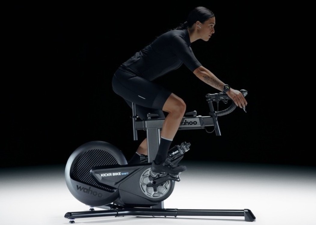 Person riding Wahoo KICKR indoor smart bike in black cycling kit, leaning forward in aerodynamic position on a studio background