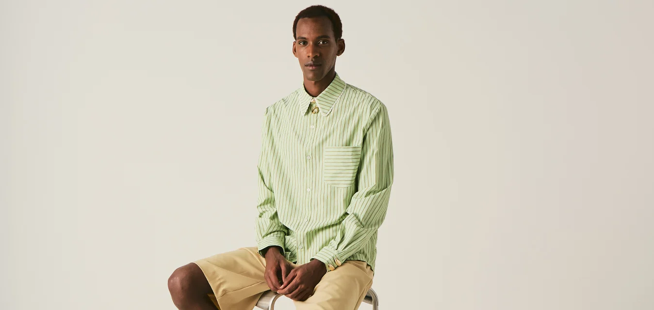 Giovane uomo seduto su uno sgabello alto in metallo, indossa una camicia verde a righe, pantaloncini beige, calzini bianchi e scarpe con stampa leopardata su uno sfondo uniforme.