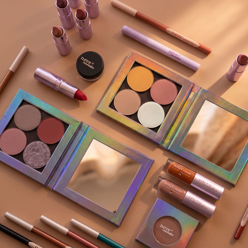 Flat lay of holographic make-up: four-pan eyeshadow palettes, lipsticks, jelly lip creams and pencils on a peach background