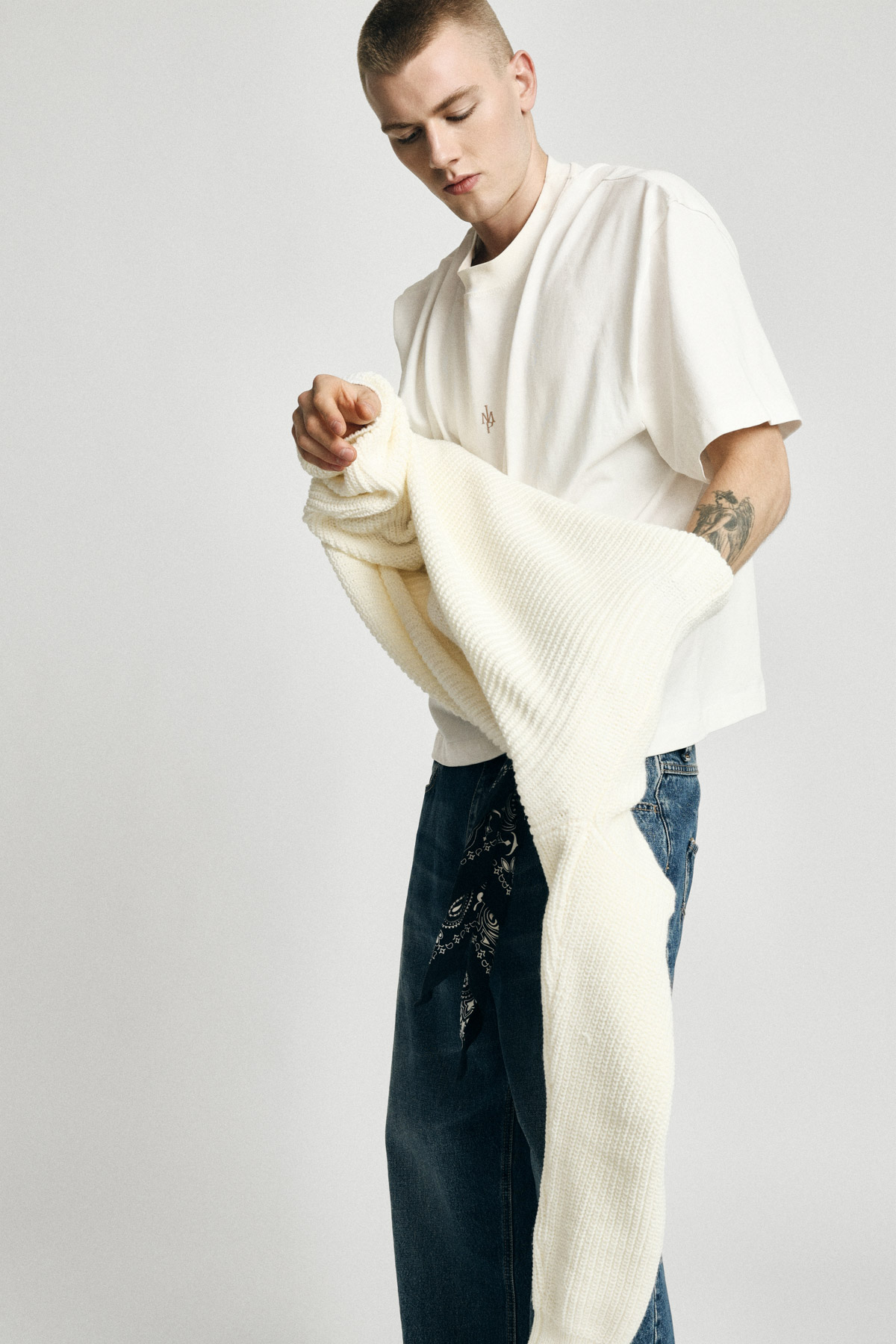 Man wearing white T-shirt and blue jeans holding a cream knitted sweater, arm tattoo visible, on a plain neutral background