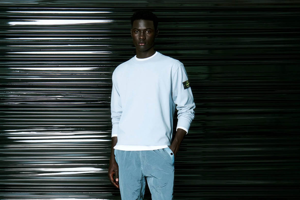 Male model in pale blue sweatshirt and light-blue trousers standing against a glossy black corrugated metal shutter