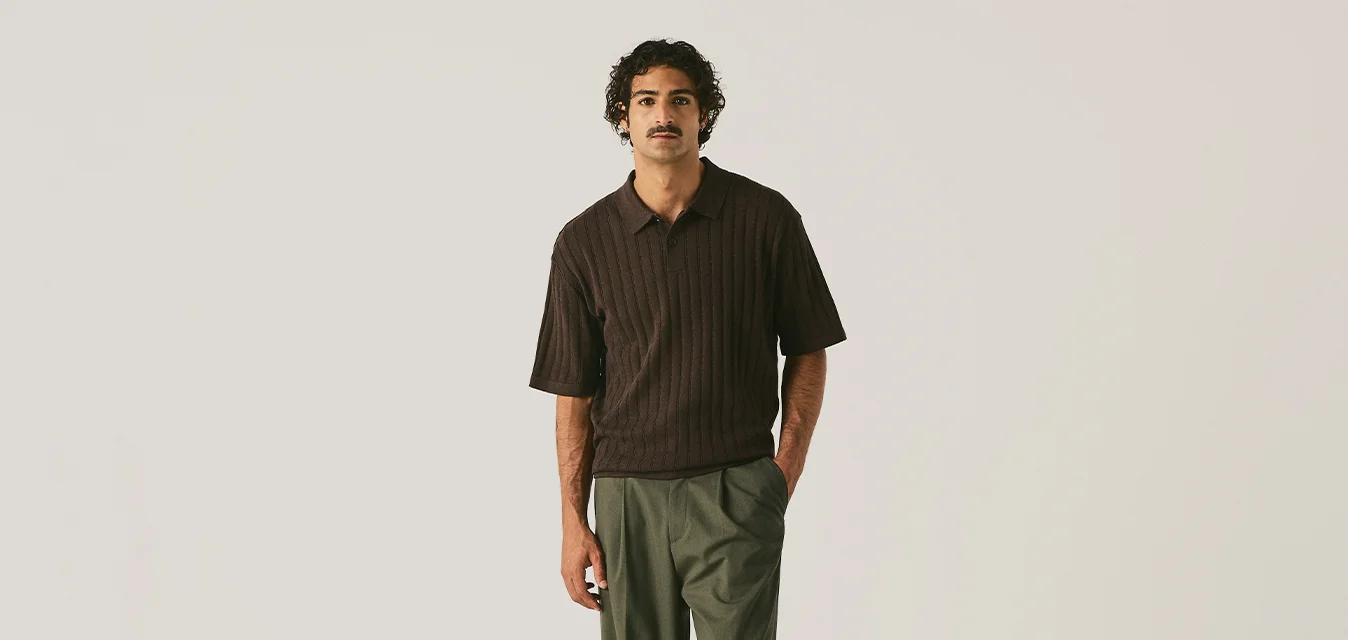 Uomo in piedi con una mano in tasca, indossa una camicia scura a maniche corte, pantaloni larghi verde scuro e scarpe marroni su uno sfondo semplice.