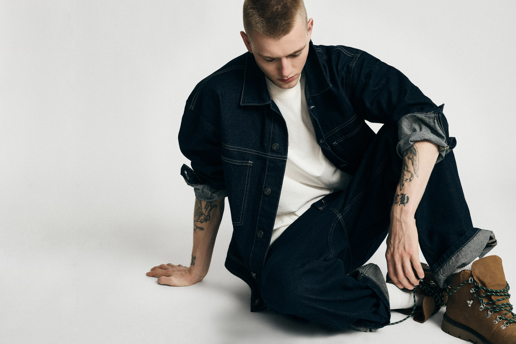 Young man model seated on studio floor wearing dark denim jacket and jeans, white T-shirt, forearm tattoos and brown hiking boots