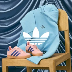 Sneaker Adidas rosa con strisce blu su una sedia di legno, accompagnati da una felpa leggera blu chiaro. Sfondo in tessuto liscio di colore blu scuro.