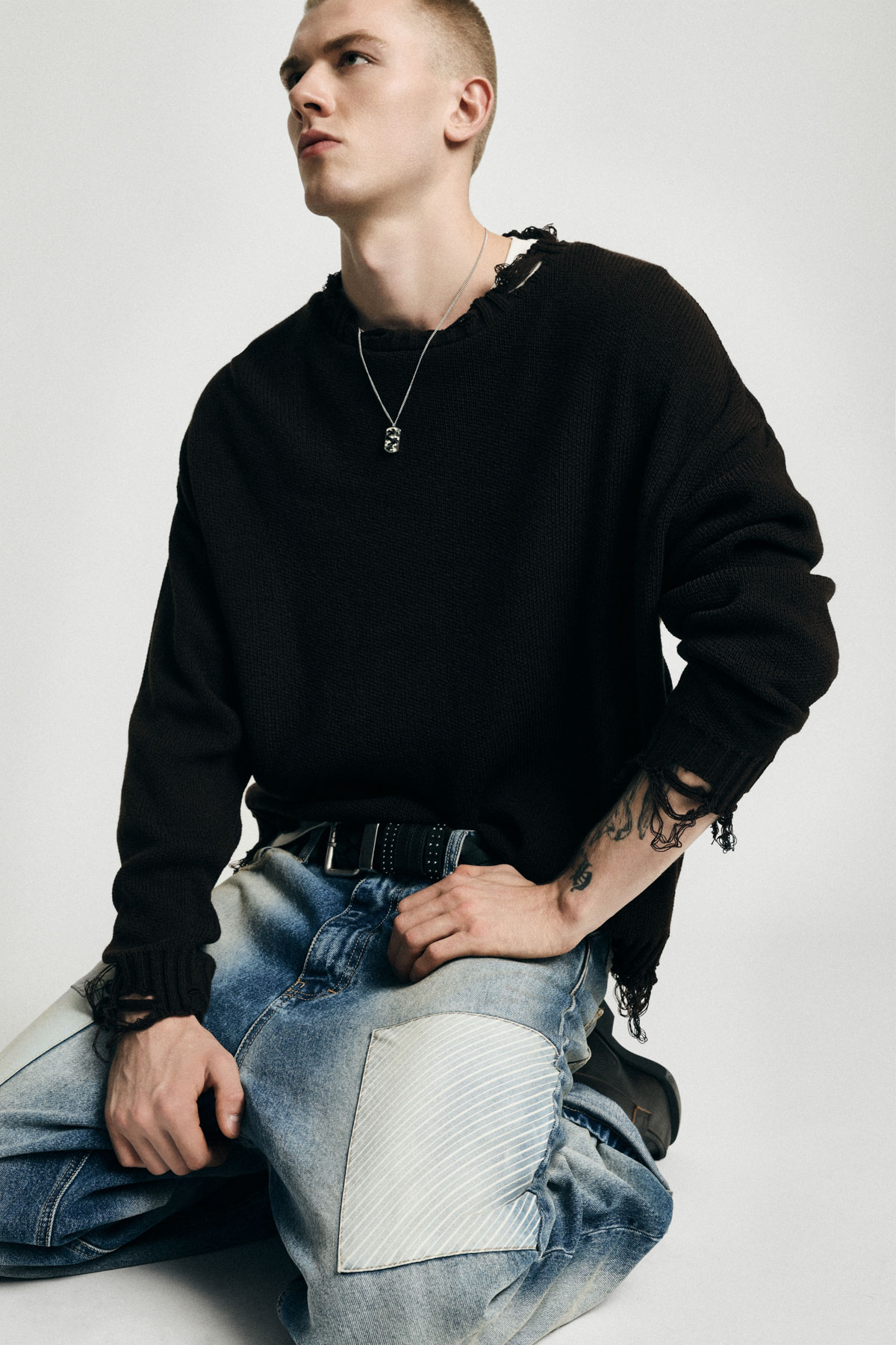 Young man seated in studio wearing black distressed sweater, pendant necklace, tattoos and patched jeans — fashion streetwear