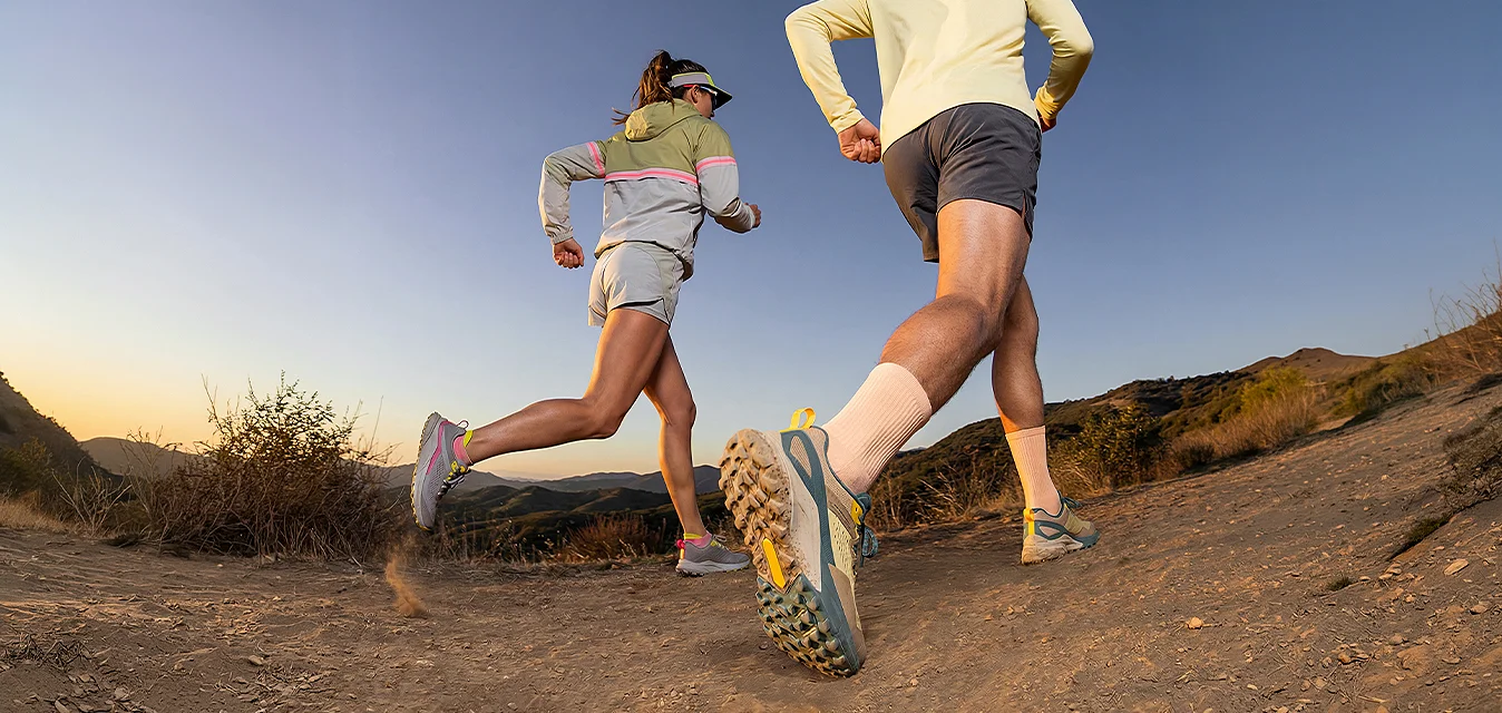 Due persone corrono su un sentiero di terra in una zona montuosa durante l'alba o il tramonto, indossando abbigliamento sportivo e scarpe da corsa.