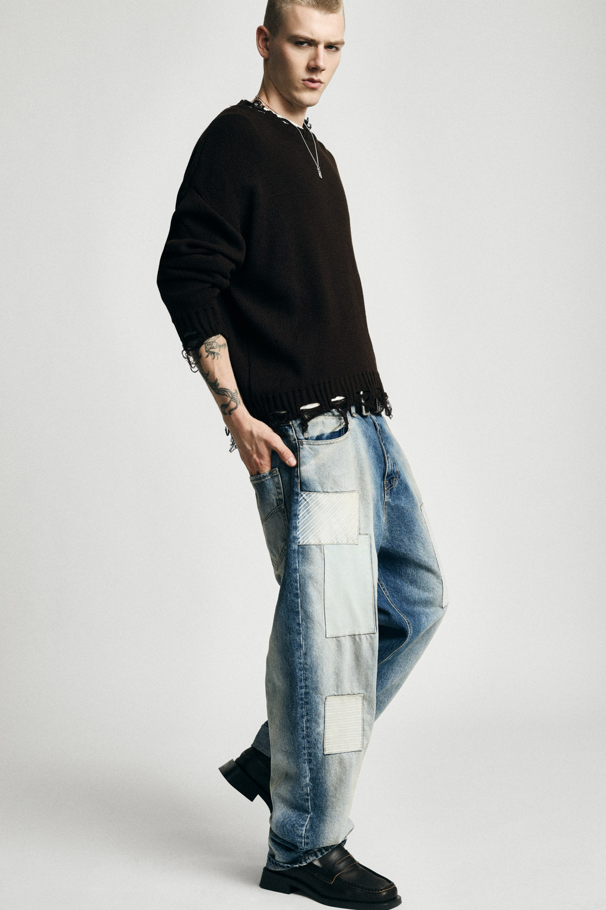 Model in black distressed sweater and patched baggy jeans, tattoos visible, standing in three-quarter pose against a plain light background