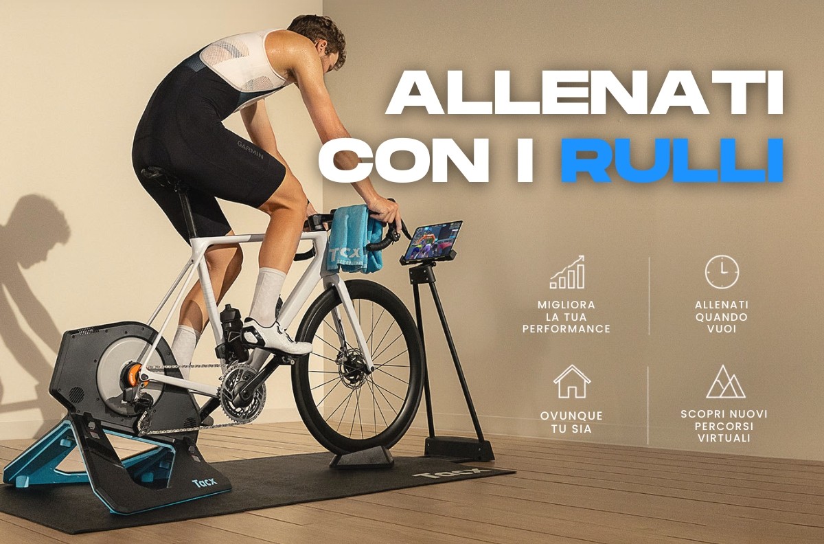 Cyclist on a road bike mounted on a smart indoor trainer with tablet display and mat, promotional text 'ALLENATI CON I RULLI'