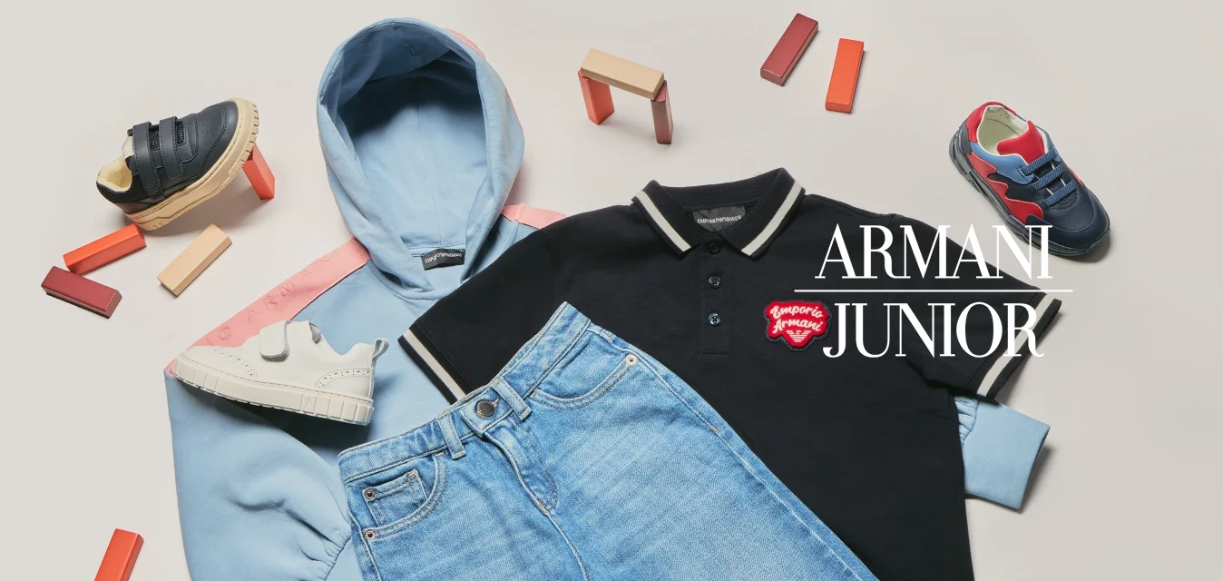 Felpa con cappuccio azzurro chiaro, polo nera con logo rosso, jeans blu e tre paia di scarpe da ginnastica colorate per bambini intorno ai vestiti su una superficie bianca.