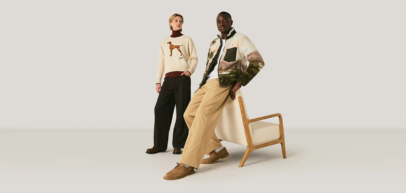 Due modelli posano in uno spazio neutro; uno è in piedi con un maglione con cane e pantaloni neri, l'altro si appoggia a una poltrona di legno indossando una giacca con fantasia e pantaloni beige.