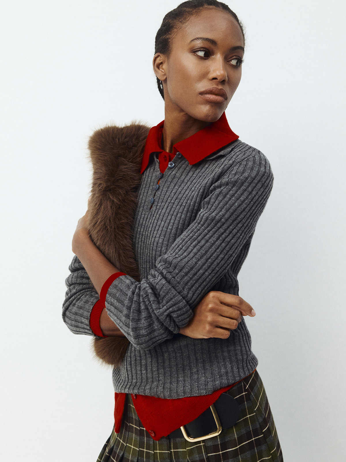 Fashion model wearing grey ribbed sweater layered over red polo shirt, tartan skirt with belt and brown fur stole, looking sideways