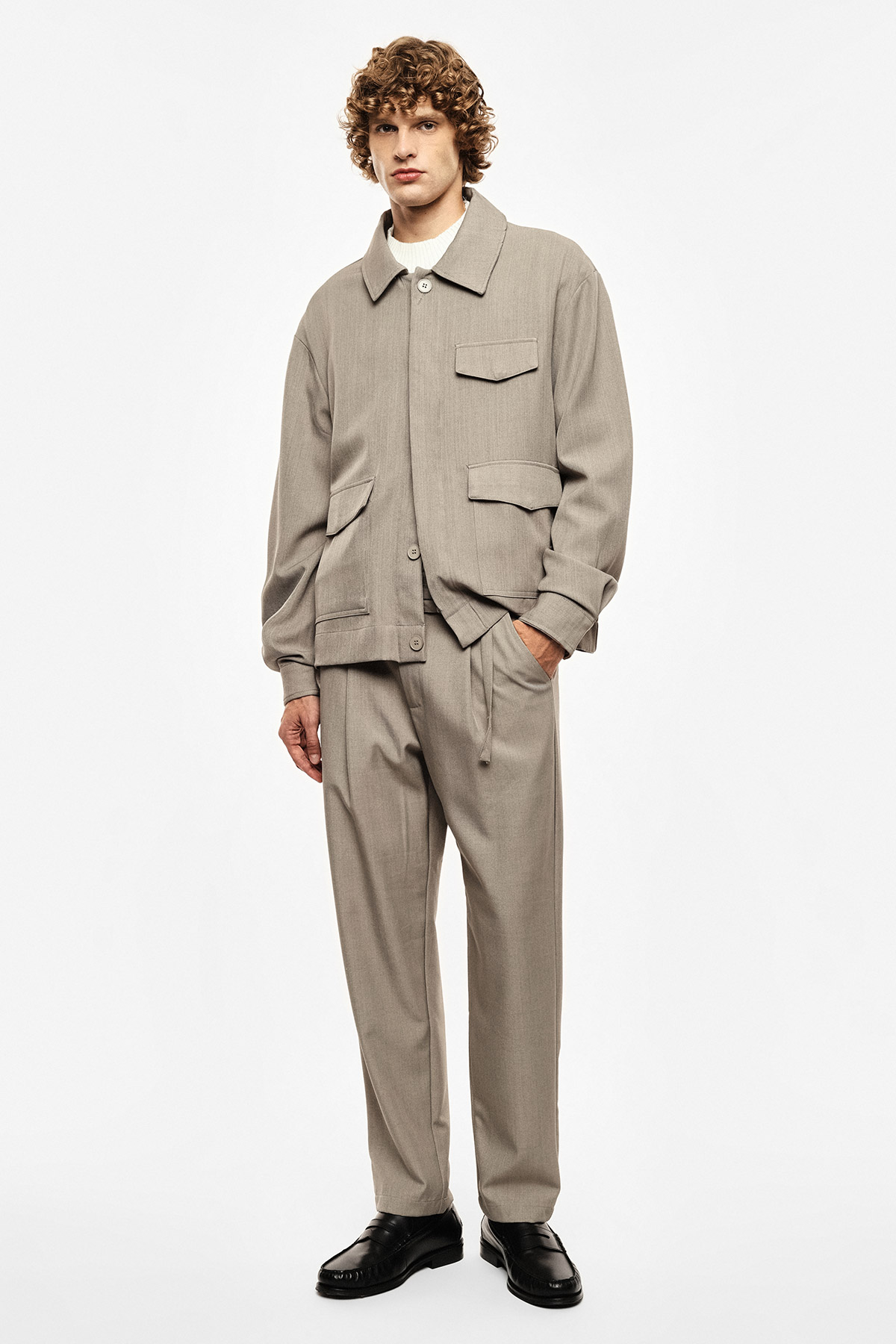 Male model wearing beige matching jacket and trousers with white T-shirt and black leather shoes, standing against plain white background