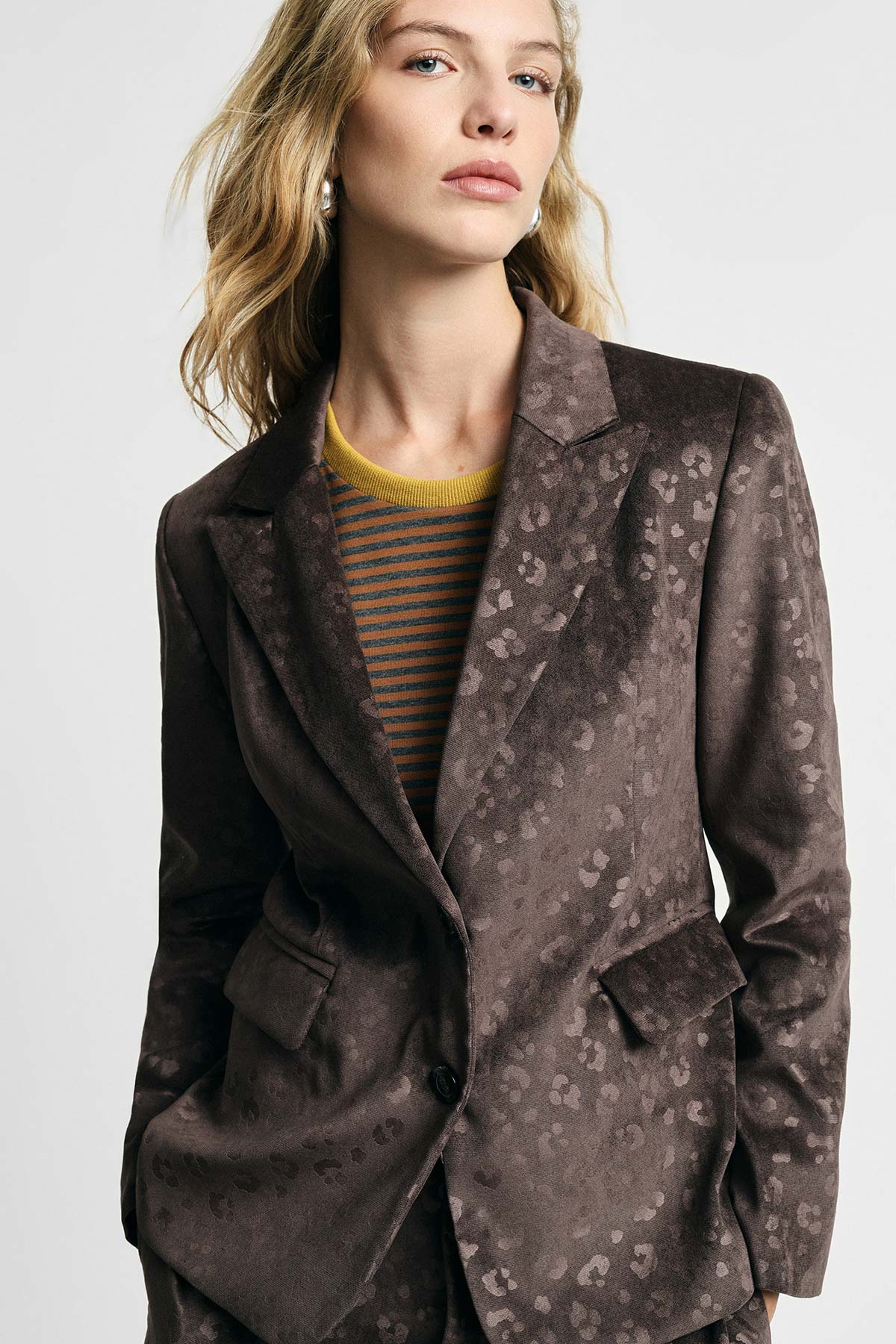 Woman wearing brown patterned velvet blazer over striped yellow and grey jumper, posing against a plain light background