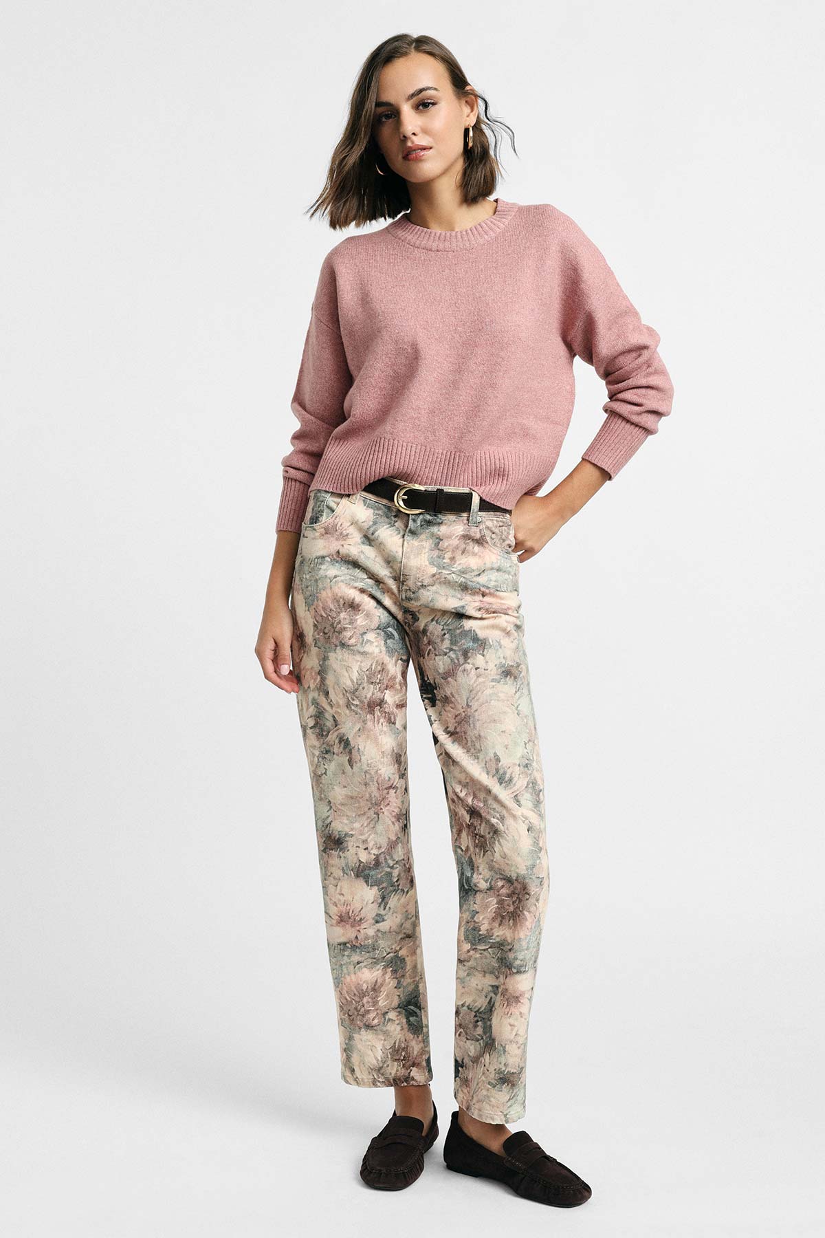 Woman in a pink knit sweater and floral print trousers with a black belt and dark loafers, standing