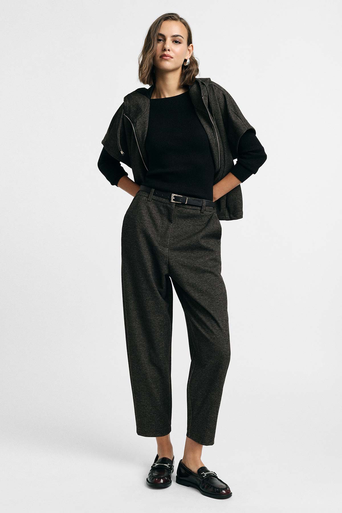 Young woman in black knit, cropped jacket, high-waisted tailored trousers and black loafers against plain white background