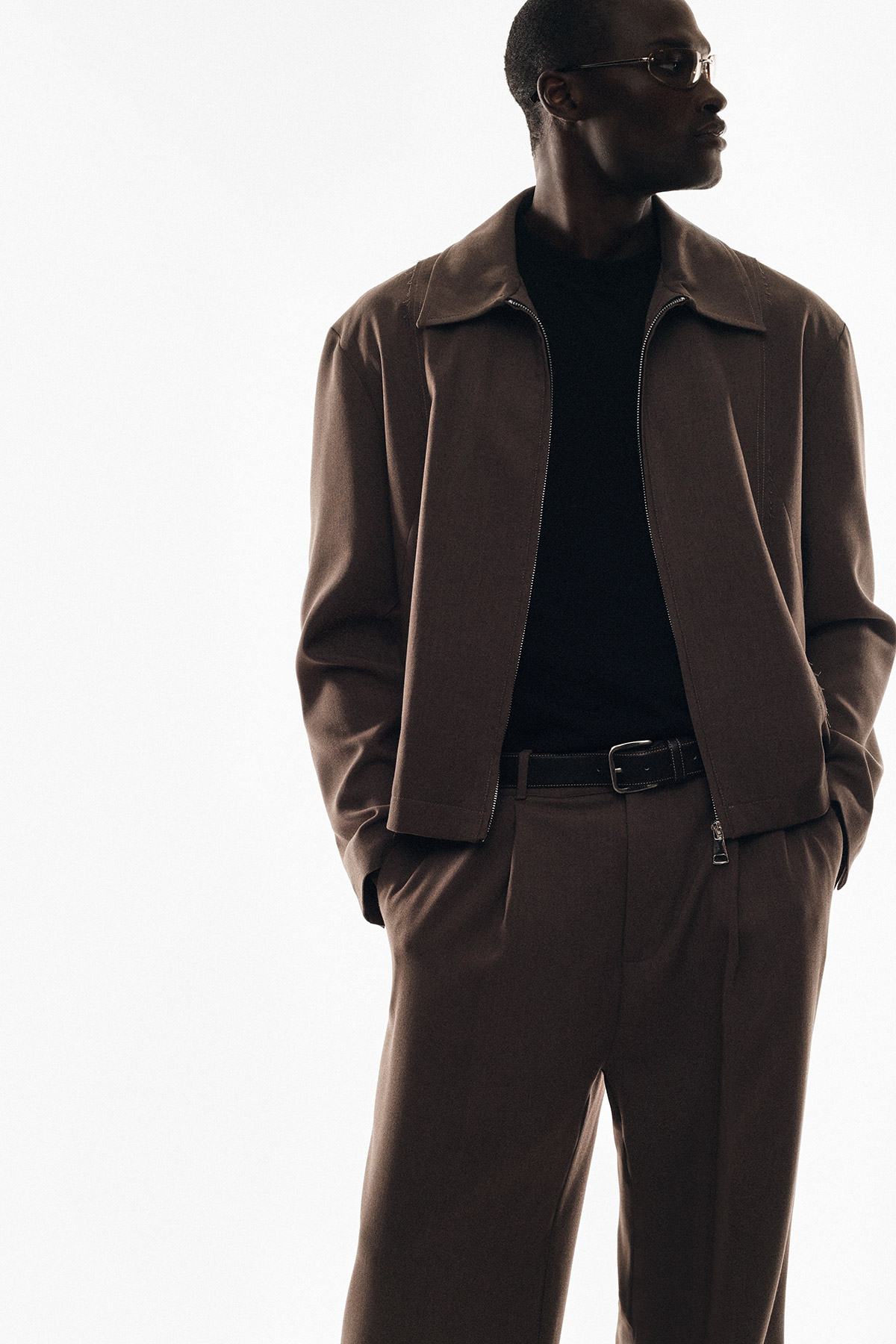 Man in brown jacket and matching trousers, black knit top and belt, wearing sunglasses with hands in pockets on white background