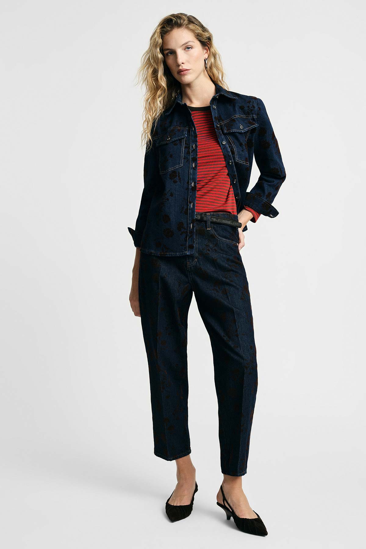 Model wearing dark patterned denim jacket and cropped matching jeans over a red striped top with black slingback heels on plain background