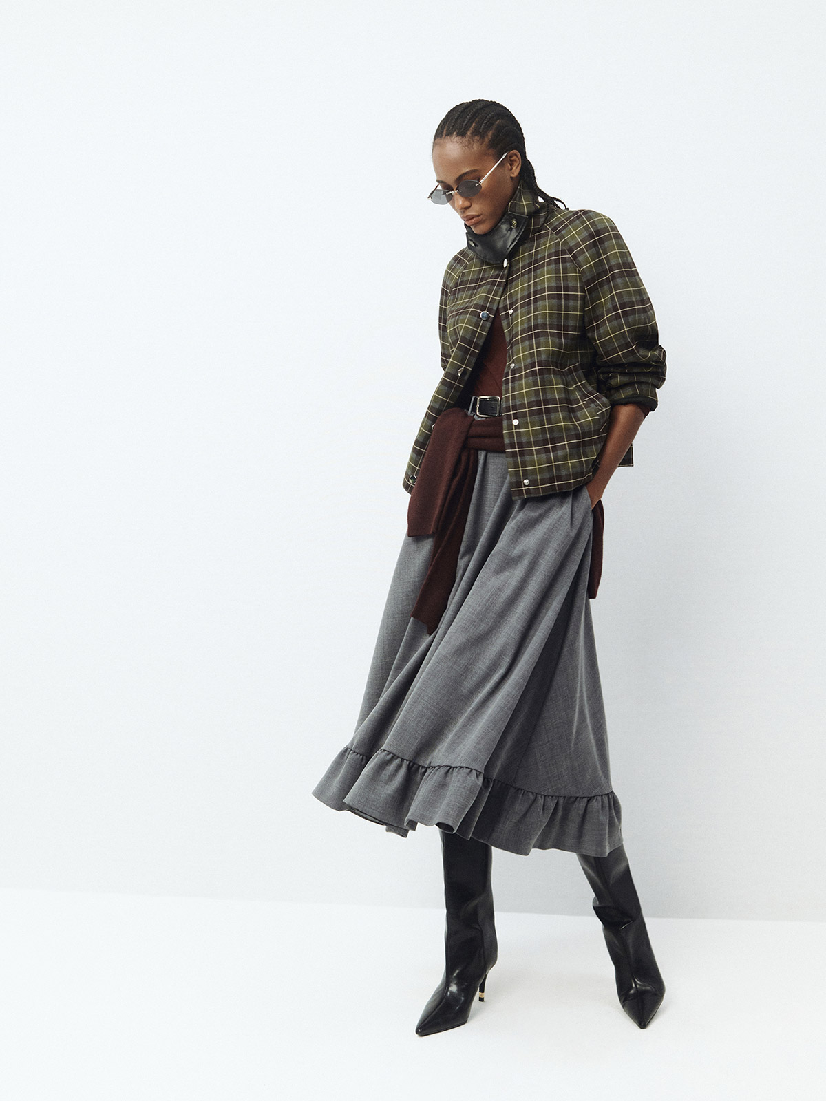 Woman in green plaid jacket, layered brown knit, long grey ruffled skirt and black heeled boots, wearing sunglasses — fashion look