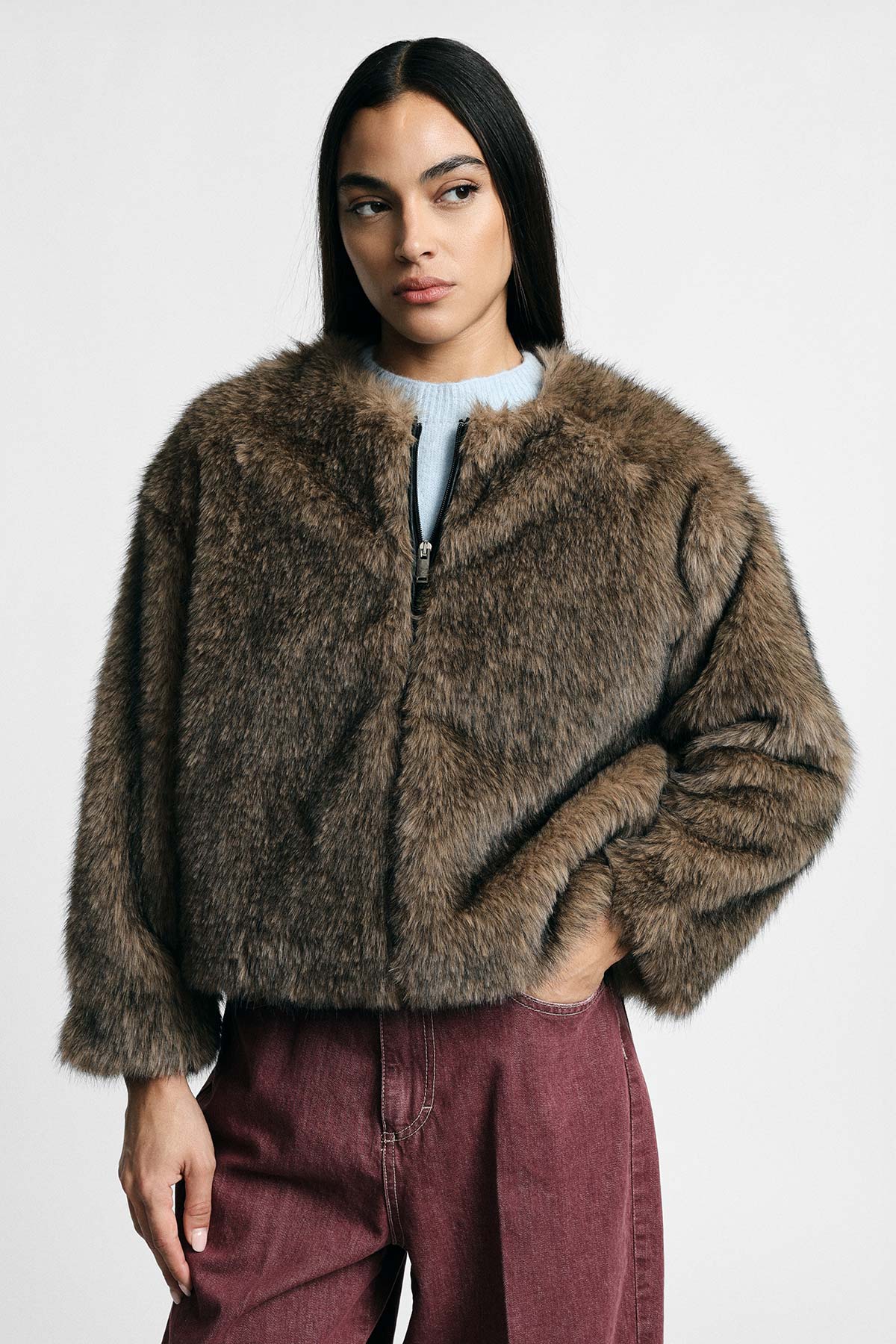 Model wearing brown faux-fur cropped jacket over light blue sweater with maroon jeans on plain grey background