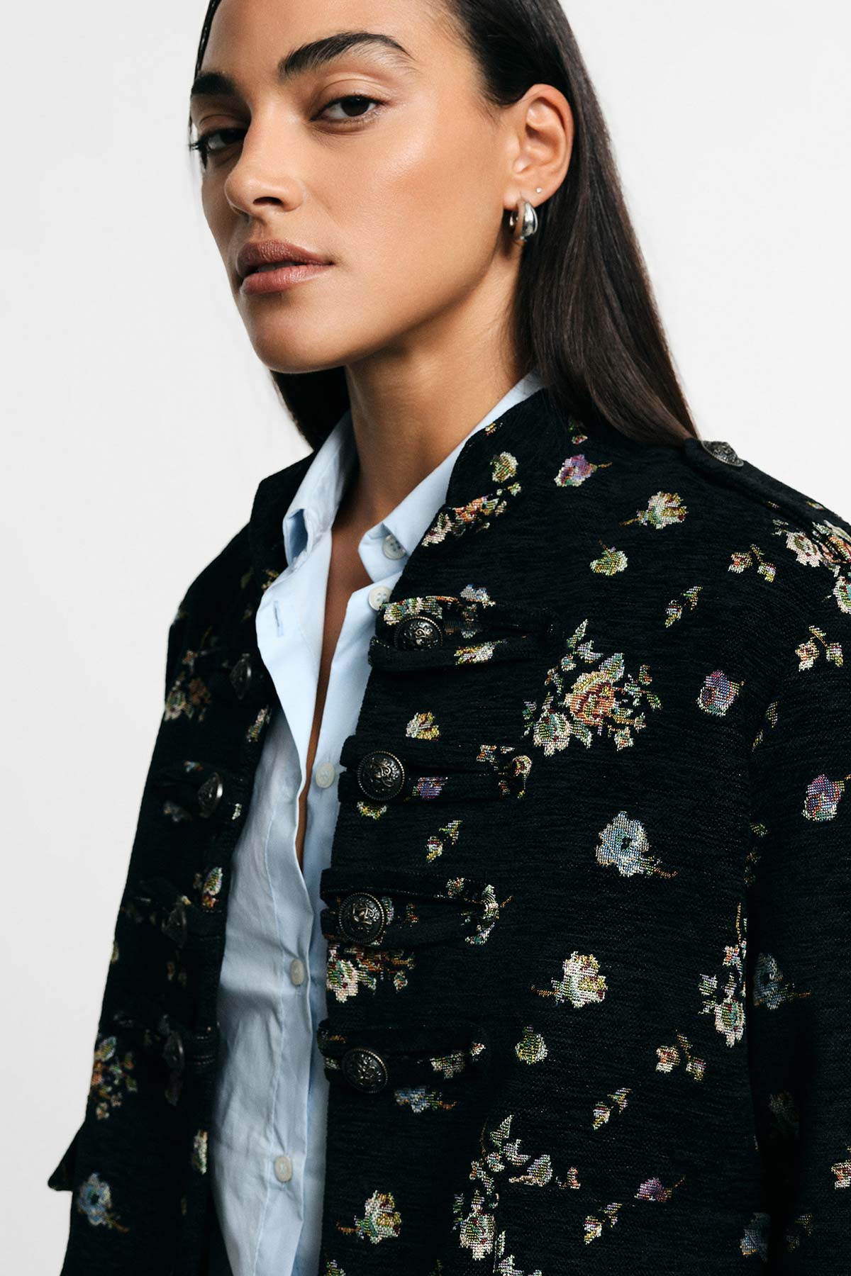 Close-up of woman wearing an embroidered black military-style jacket over a light blue shirt with a silver hoop earring