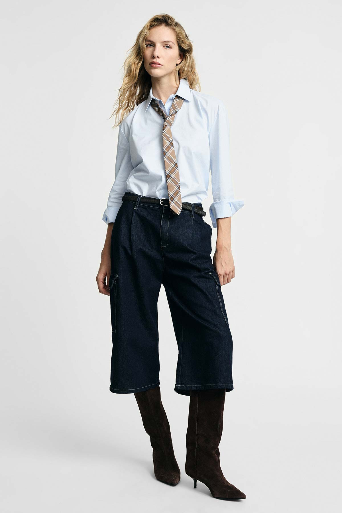 Blonde woman in pale blue shirt with plaid tie, cropped dark denim trousers and brown knee‑high suede boots on white