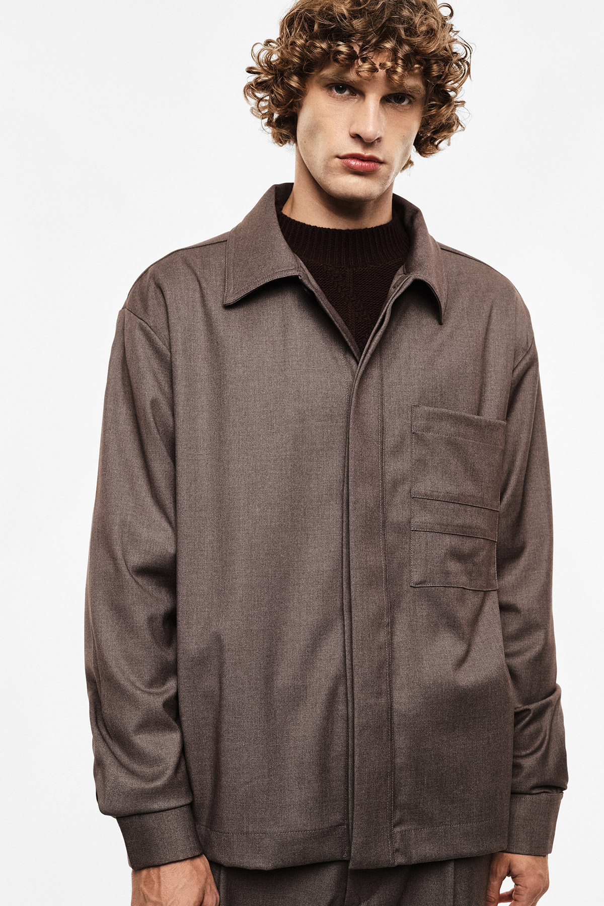 Curly‑haired man wearing a brown wool overshirt over a dark sweater, standing front‑on against a plain white background