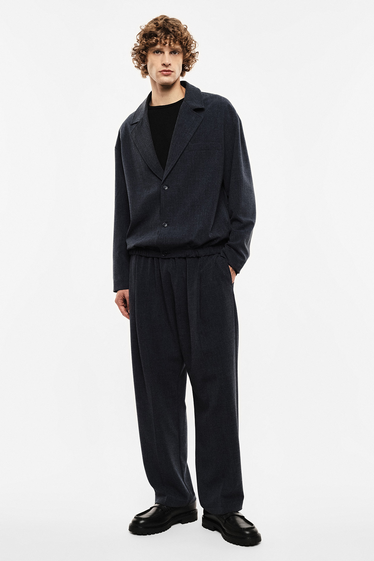 Man wearing navy relaxed two-piece suit with elastic-waist trousers, black jumper and loafers on white background