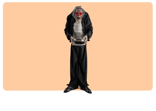 Creepy Halloween ghoul prop with skull-like face and glowing red eyes in tattered black suit holding a silver bowl on peach background