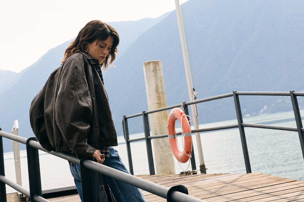Woman in a brown leather jacket leaning on a railing at a wooden lakeside pier with a lifebuoy and mountains
