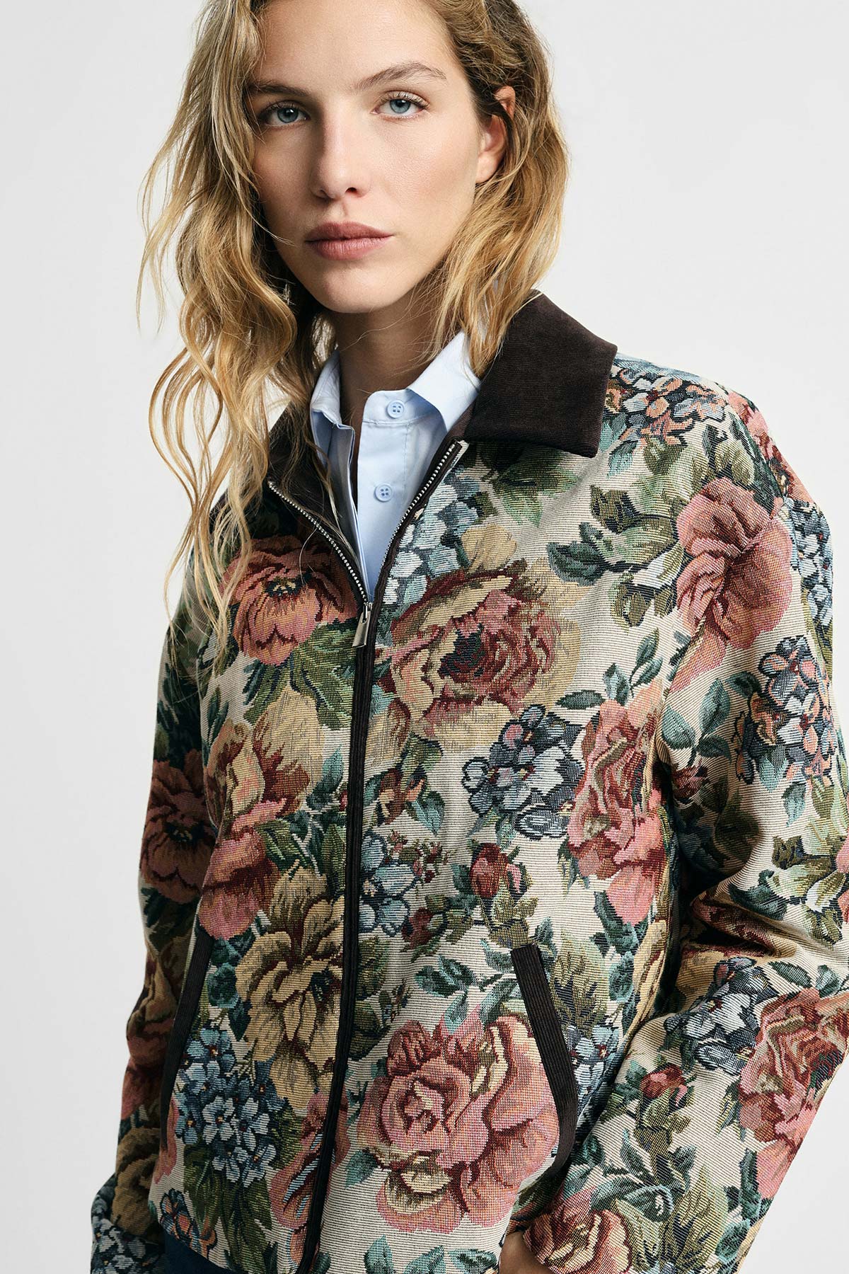 Woman wearing floral tapestry bomber jacket with corduroy collar over blue shirt, close-up portrait on neutral background