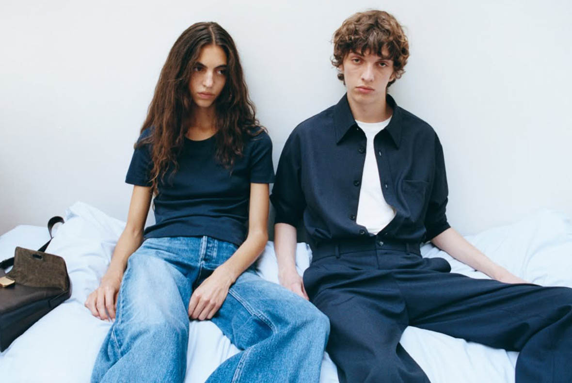 Two young adults sitting on a bed in a minimalist room, wearing casual navy tops and denim jeans — relaxed fashion portrait