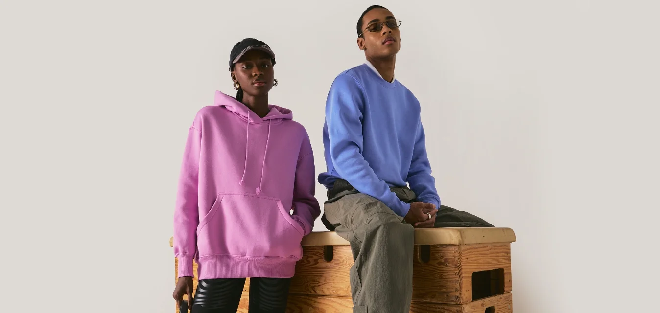 Donna con felpa rosa con cappuccio e pantaloncini neri accanto a uomo con felpa blu e pantaloni cargo seduto su una scatola di legno per ginnastica.