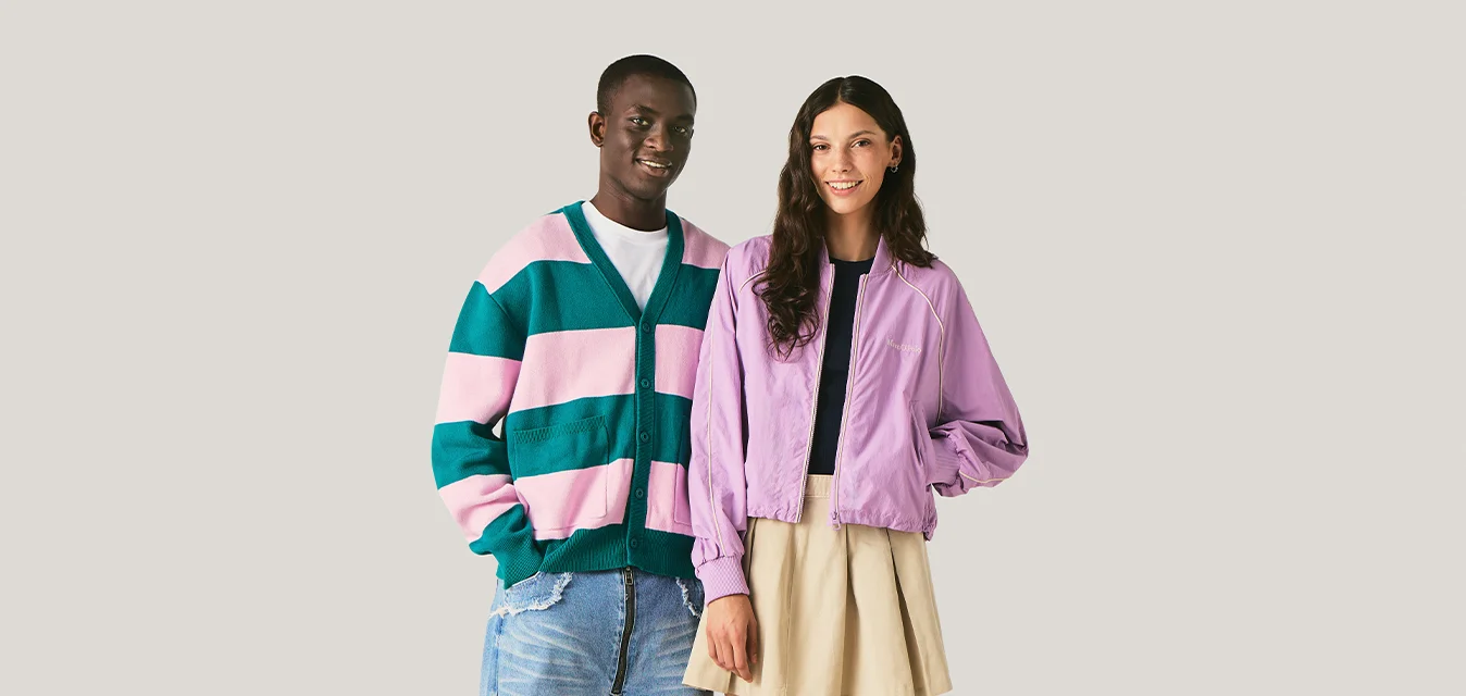 Giovane uomo sorridente con maglione a righe verdi e rosa in piedi accanto a giovane donna sorridente con giacca viola e gonna beige su sfondo neutro.