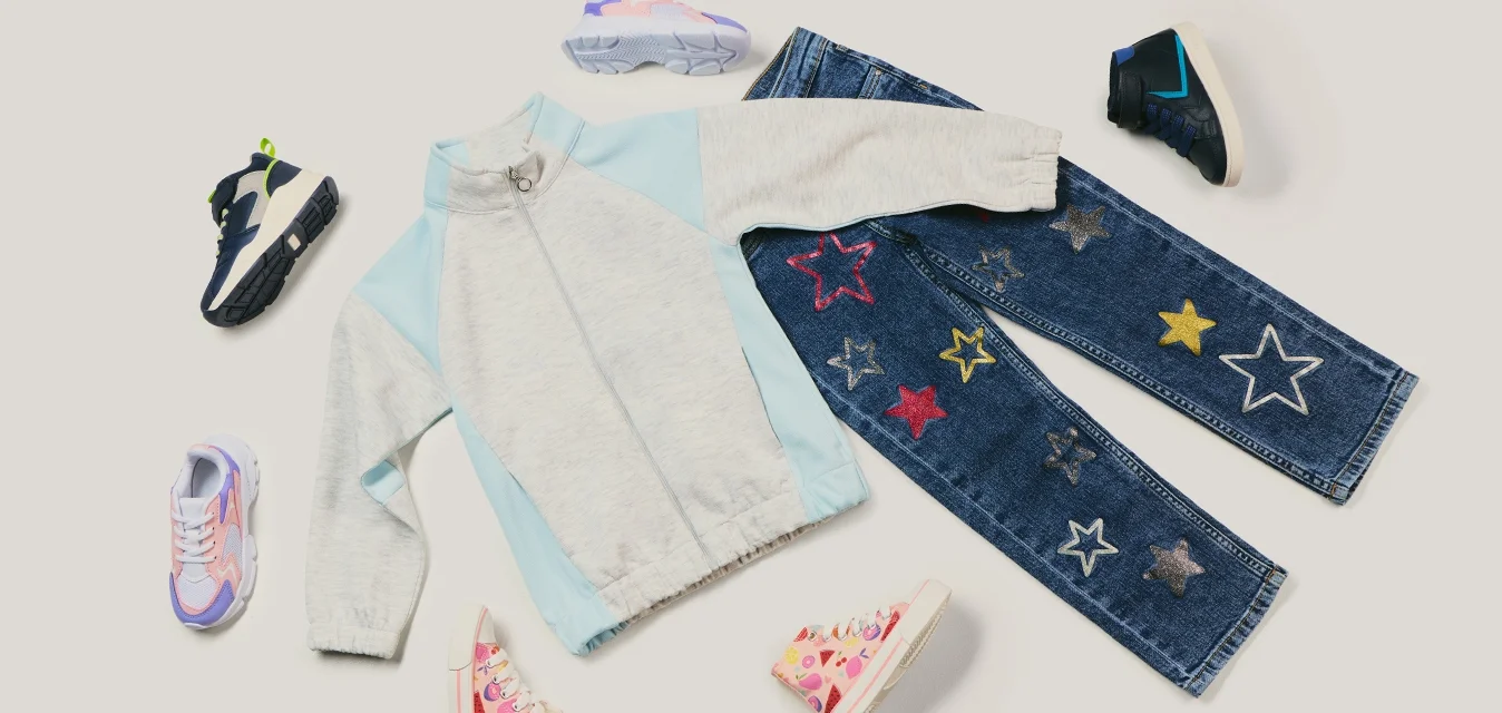 Giacca leggera per bambini azzurro chiaro e grigio con zip, abbinata a jeans blu decorati con toppe a forma di stelle colorate e circondati da cinque paia di scarpe da ginnastica diverse.