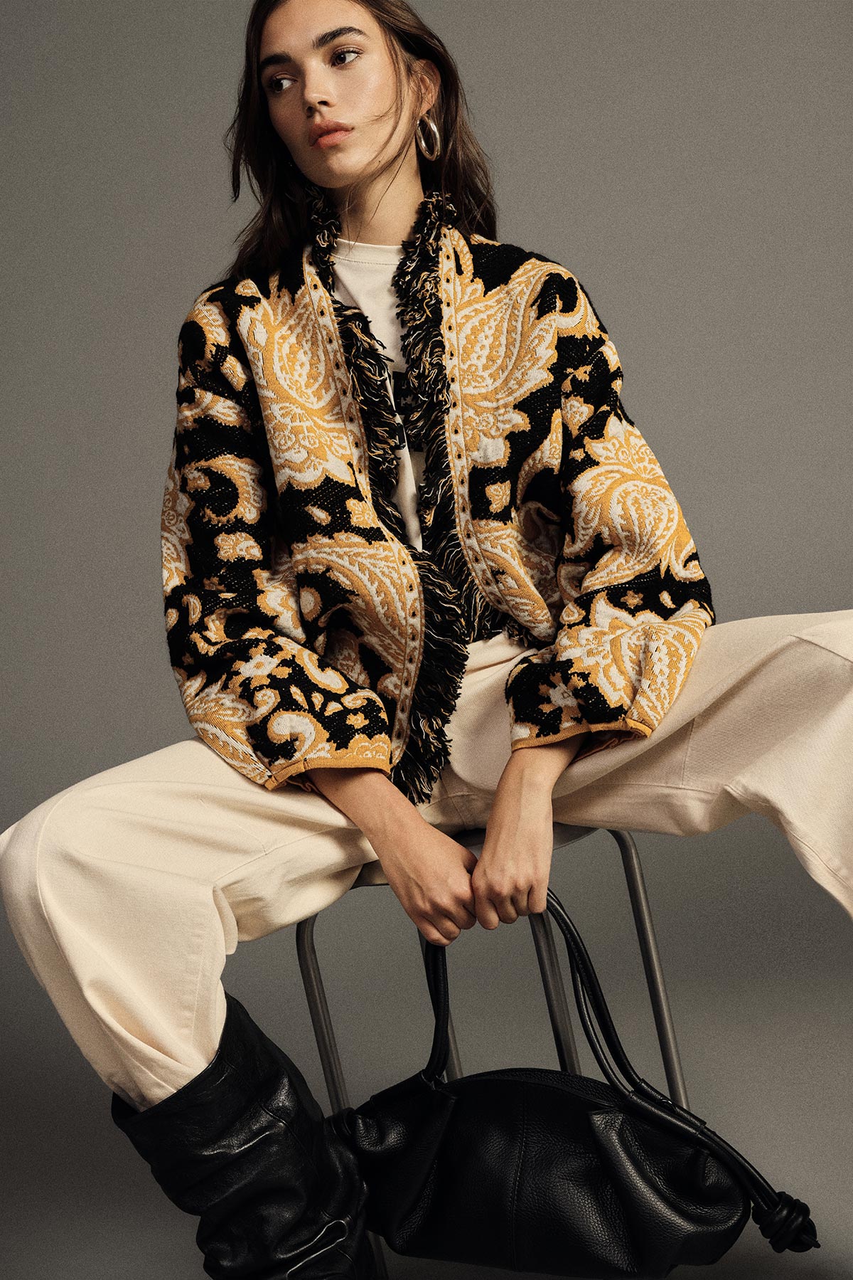 Woman seated wearing a black and gold paisley jacket, cream trousers and black boots, holding a slouchy black leather handbag