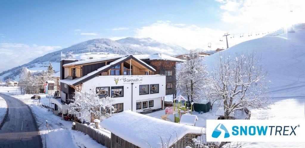 Snow-covered alpine guesthouse with playground, ski slopes and chairlift under blue sky, SnowTrex logo