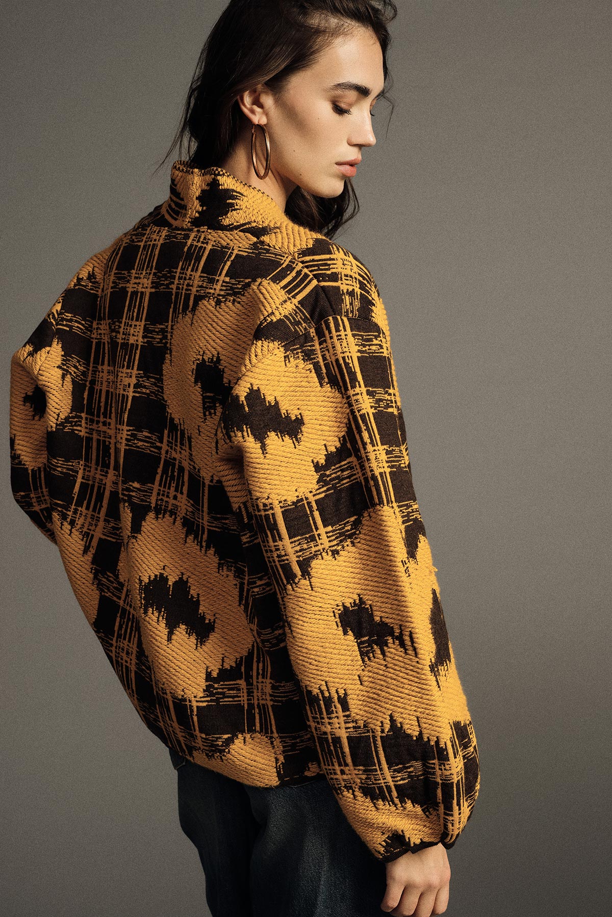 Model wearing a yellow and black patterned knit jacket with hoop earrings, side pose against a plain grey background, fashion knitwear