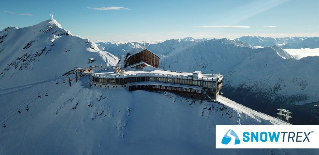 Aerial view of a snow-covered alpine summit station and restaurant with ski lifts on a mountain ridge, SnowTrex logo