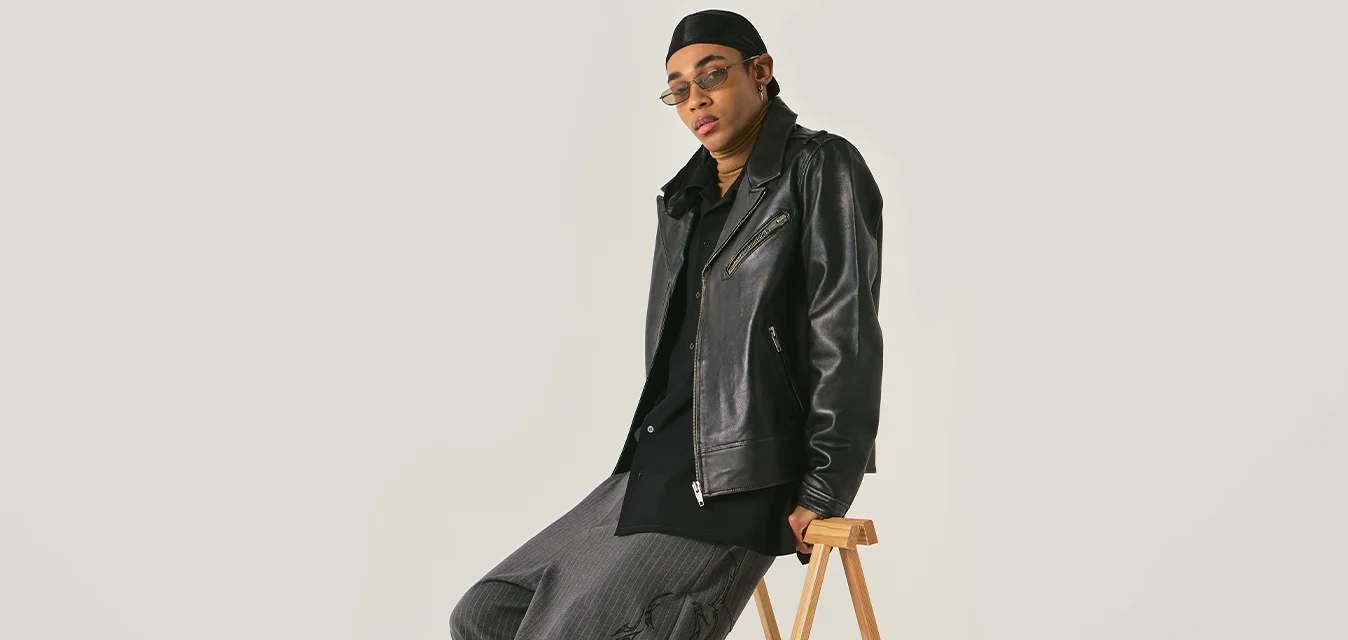 Persona con giacca di pelle nera, pantaloni grigi larghi con motivo, occhiali da sole e cappello, appoggiata a uno sgabello pieghevole in legno davanti a uno sfondo semplice.