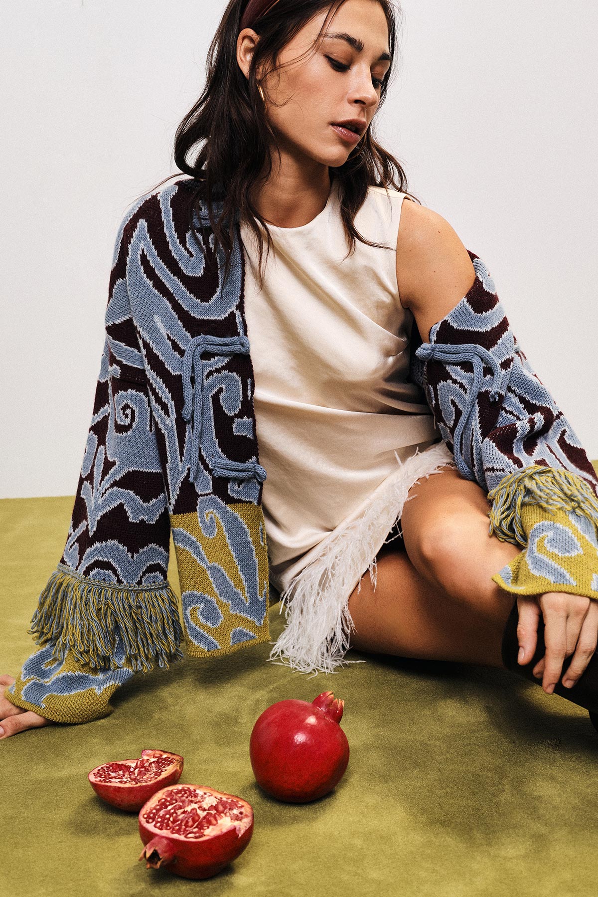 Young woman seated in patterned fringed cardigan and cream dress with pomegranates on green surface, fashion editorial image