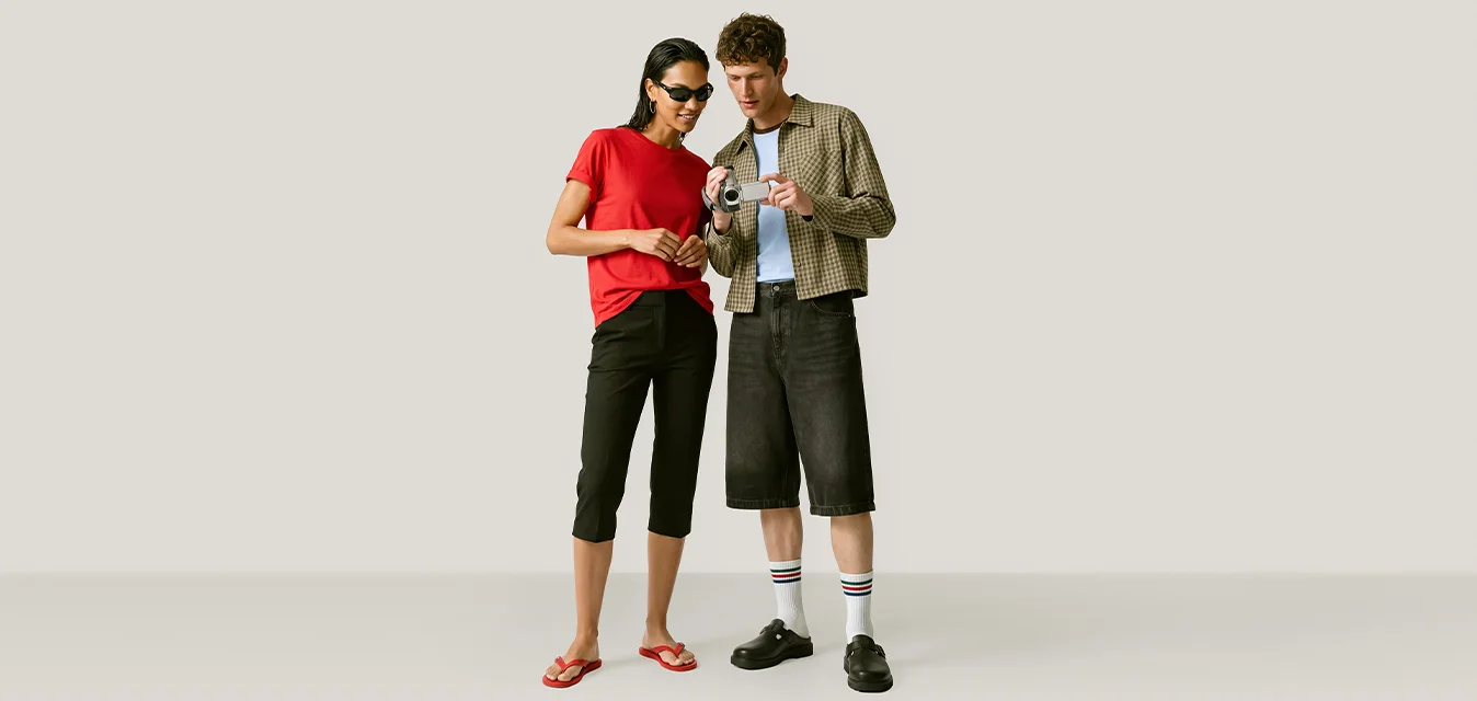 Una donna con una camicia rossa e pantaloni neri e un uomo con una giacca a quadri e pantaloncini neri guardano insieme una fotocamera digitale.