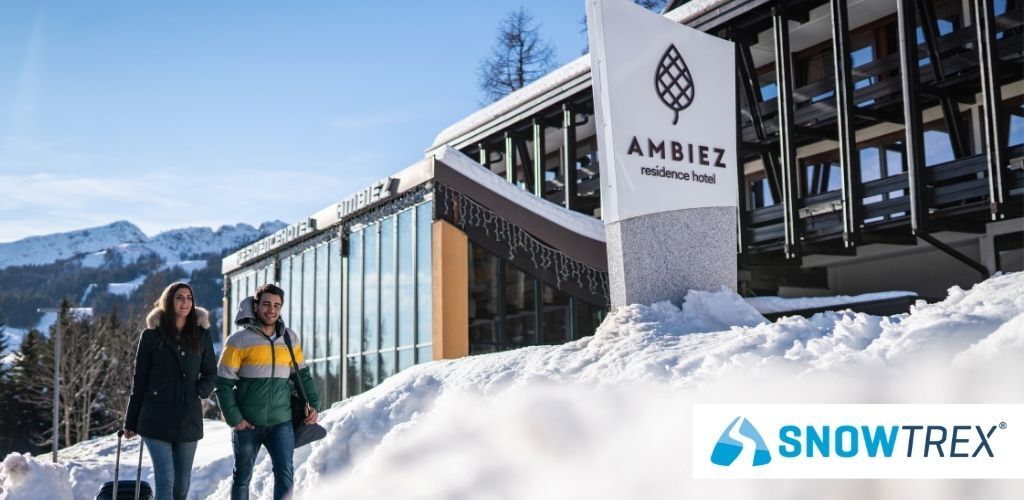 Two travellers walking with luggage past the Ambiez Residence Hotel sign in a snowy alpine resort with SnowTrex logo