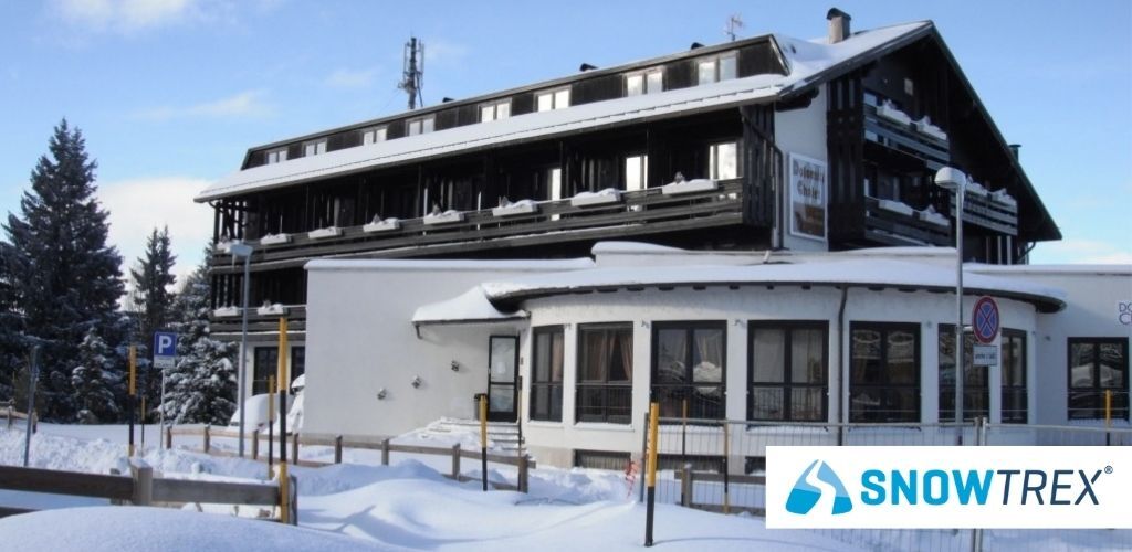 Snow-covered chalet-style hotel with fenced snowy path and SnowTrex logo in bottom-right corner