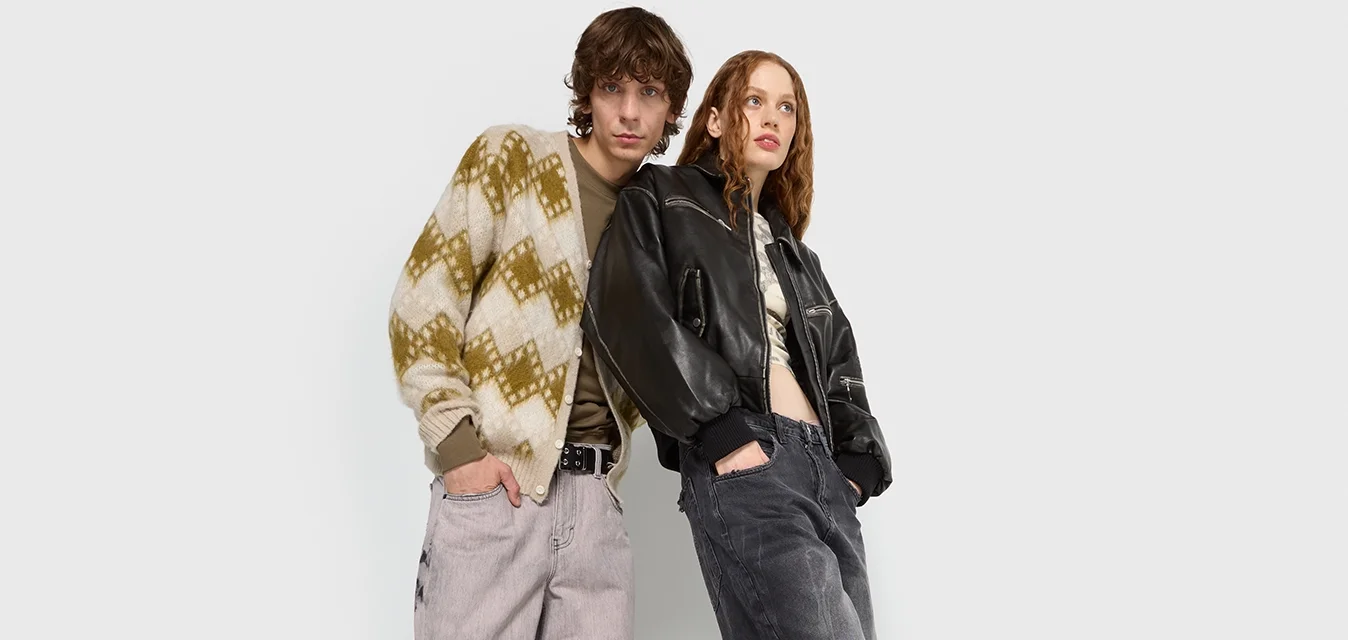 Due modelli si trovano davanti a uno sfondo neutro. Uno indossa un maglione a fantasia e pantaloni larghi di colore chiaro; l'altro indossa una giacca di pelle nera e pantaloni larghi scuri.